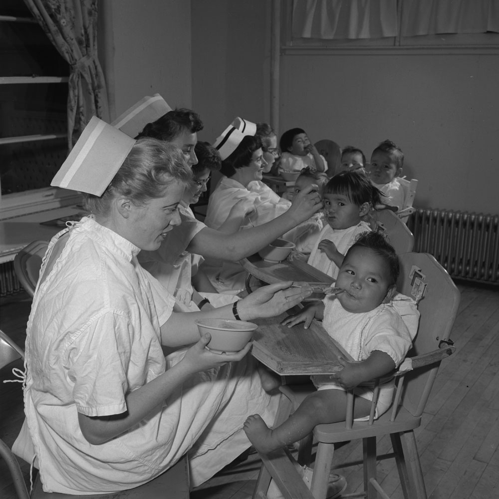 nurses in caps feed babies in high chairs