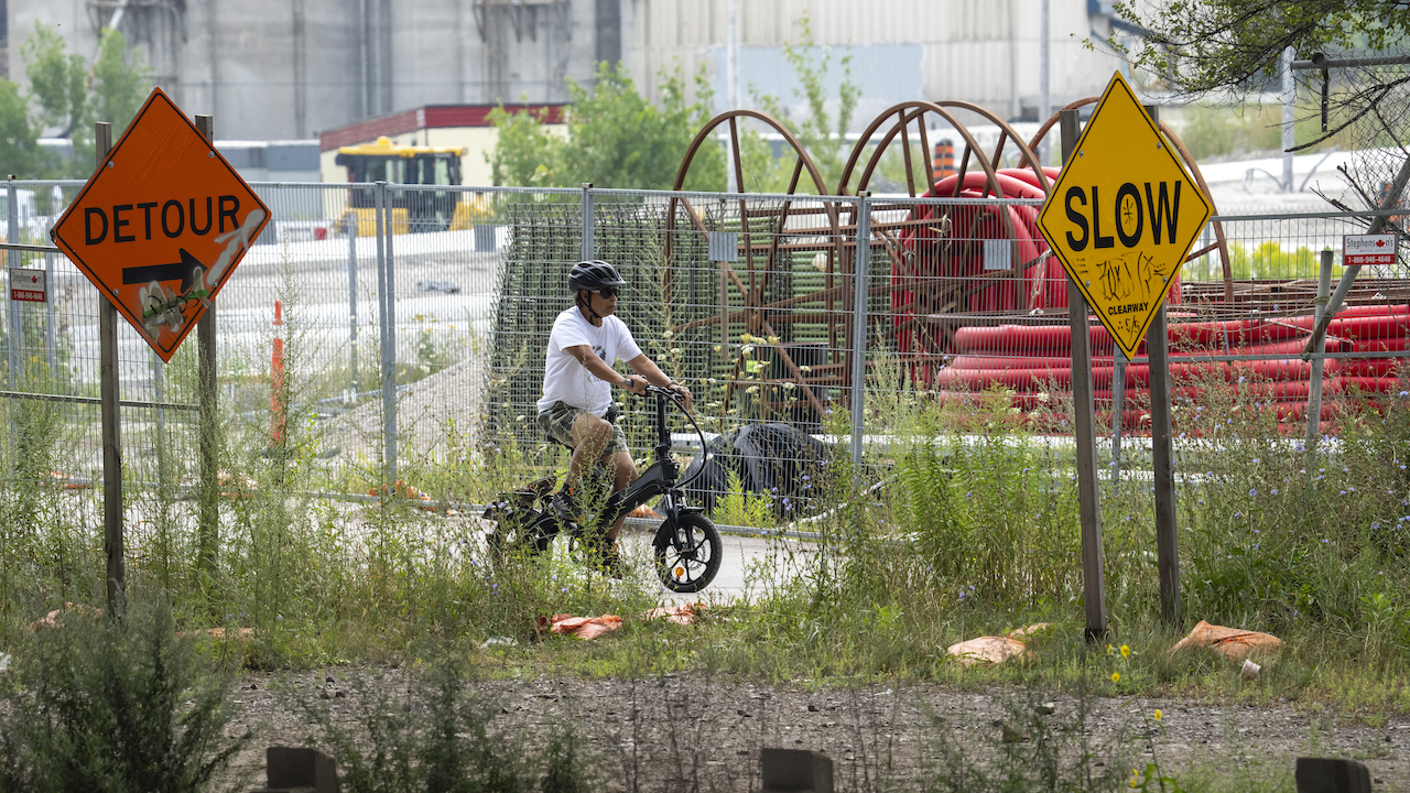 cyclist on a path in front of construction equipment and between signs reading "Detour" and "Slow"