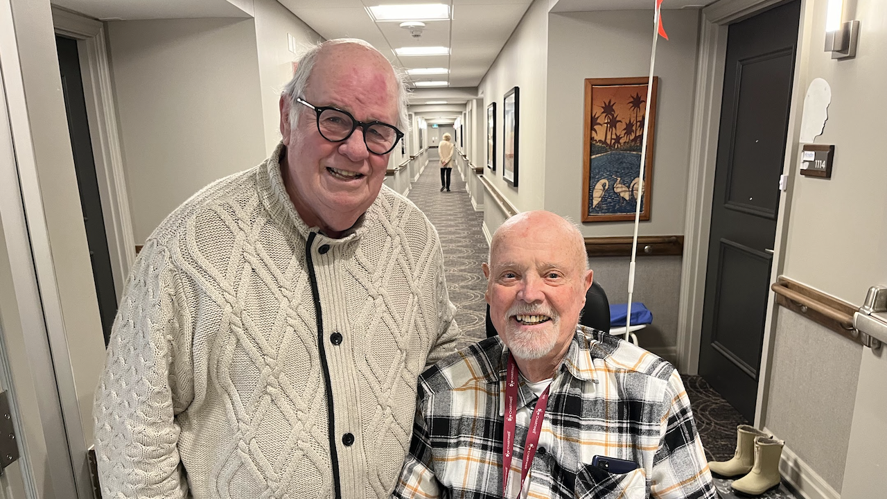 two smiling men, one standing and one seated, in a hallway