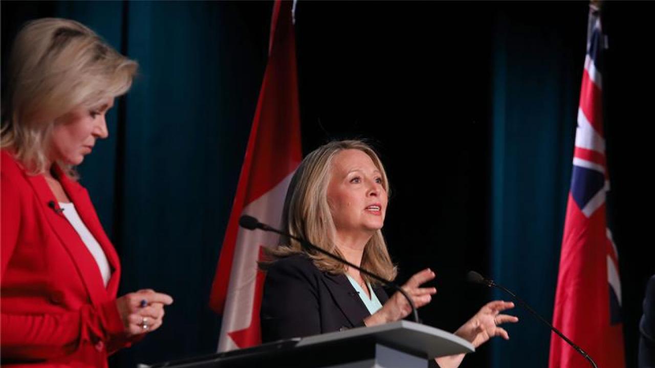 blonde woman in red blazer listens as blonde woman in black blazer speaks and gestures