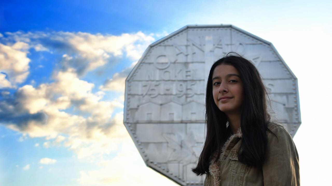 smiling long-haired young woman stands in front of giant nickel under a blue sky