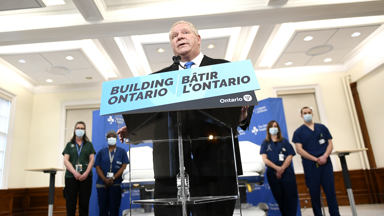 suited man speaks behind a lectern ("Building Ontario" while masked health-care workers look on