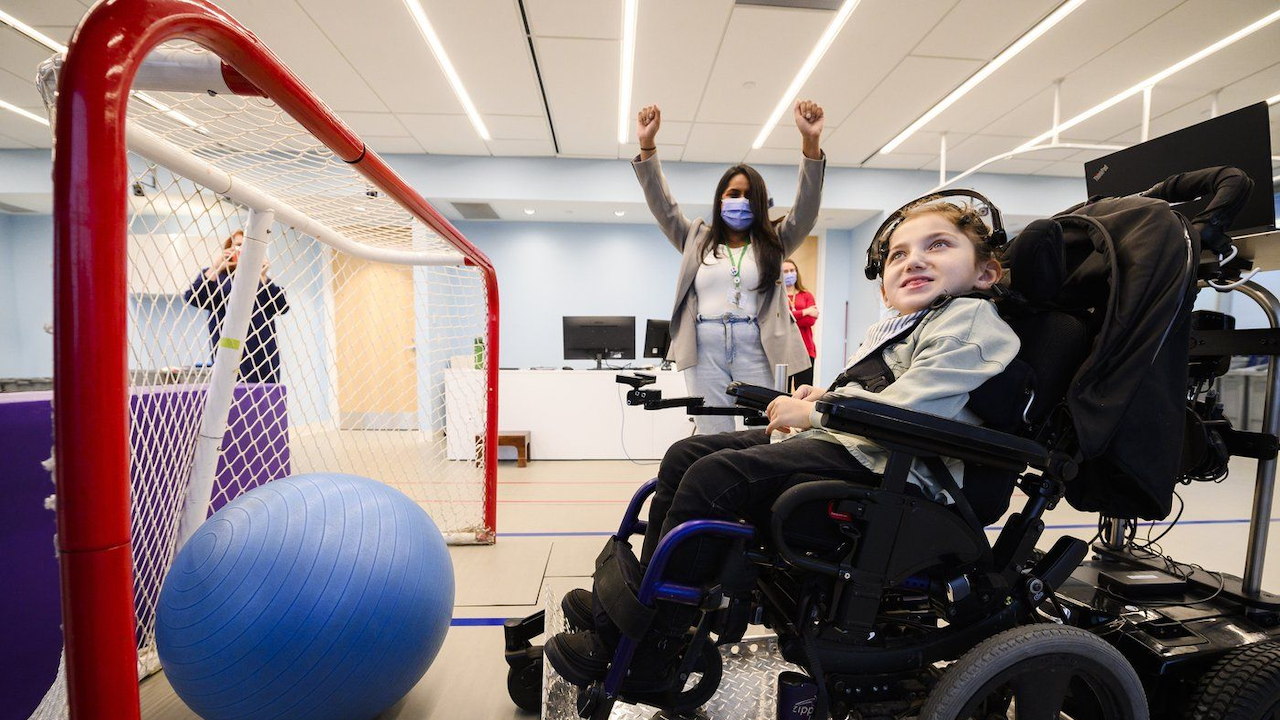 child in wheelchair sits in front of ball in net while woman behind them raises both arms in victory
