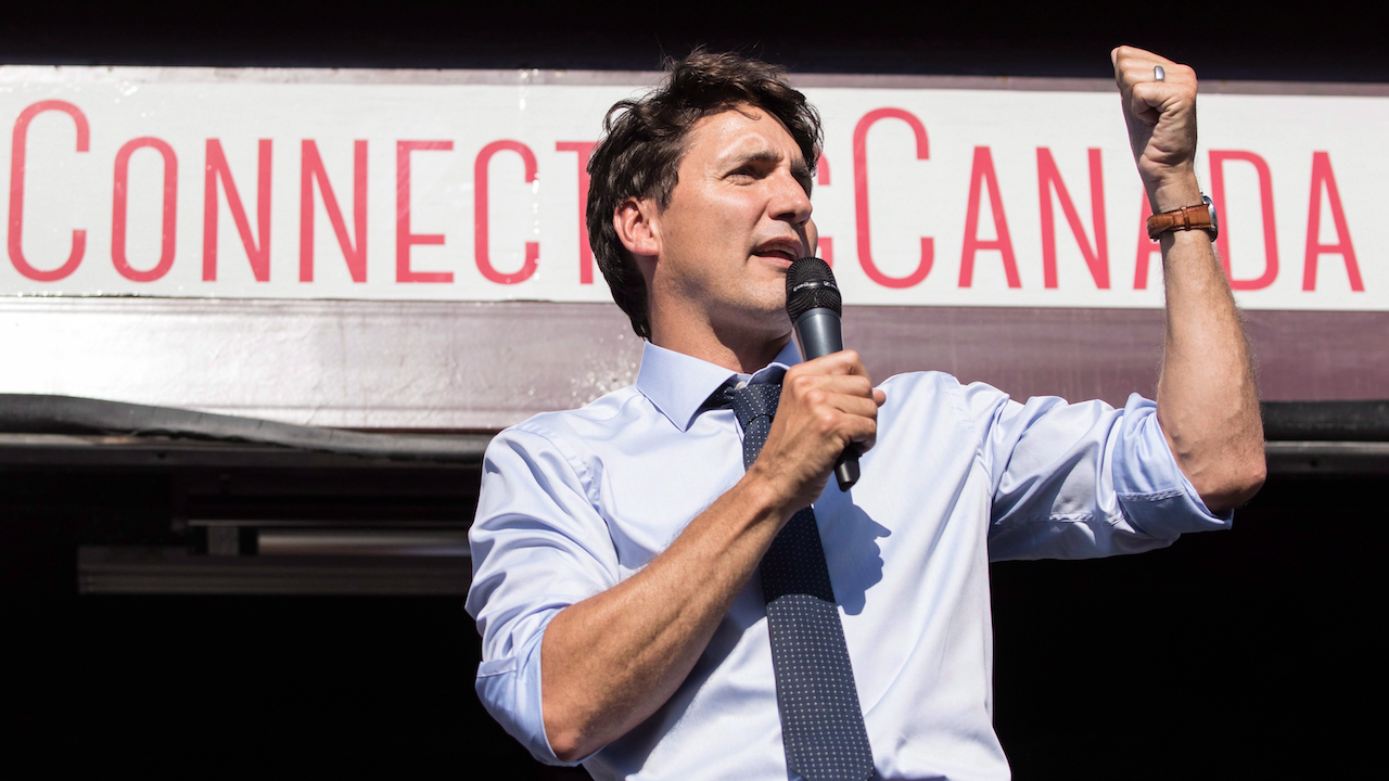 man in button-up shirt and tie speaks into a microphone with one hand raised in front of a sign reading "Connecting Canada"